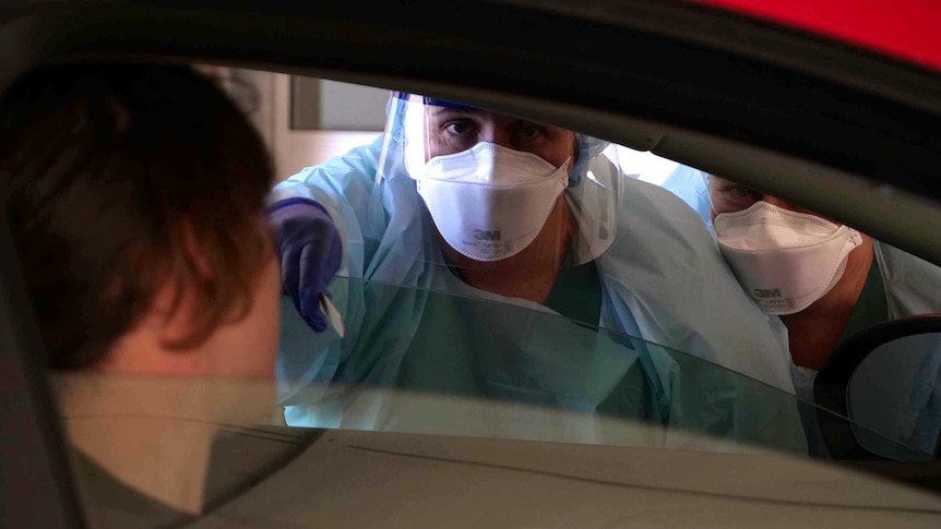 A medical professional dressed in protective clothing leans through the window of a car to test people inside.