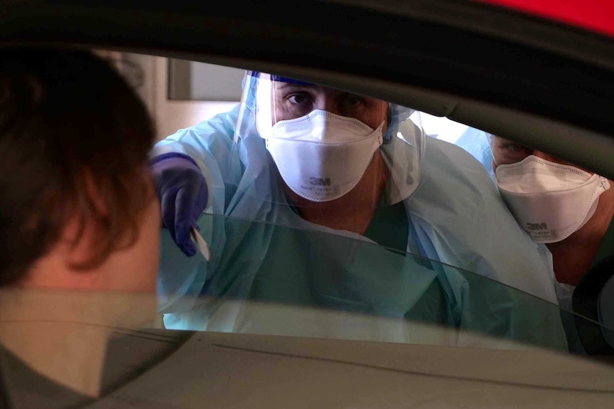 A medical professional dressed in protective clothing leans through the window of a car to test people inside.