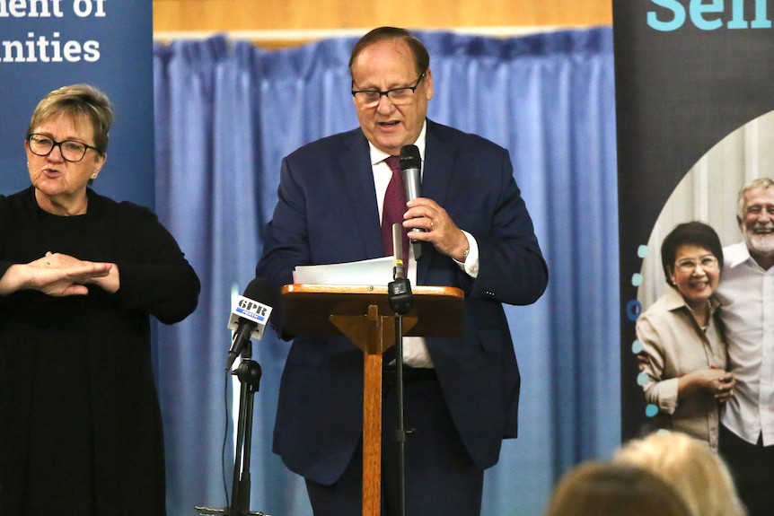 Don Punch at a podium holding a microphone, wearing a blue suit and red tie and giving a speech, next to an AUSLAN interpreter.