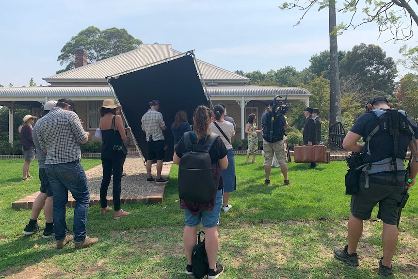 Crew with cameras and light board filming Annabel Crabb talking to family outside a Federation-era house.