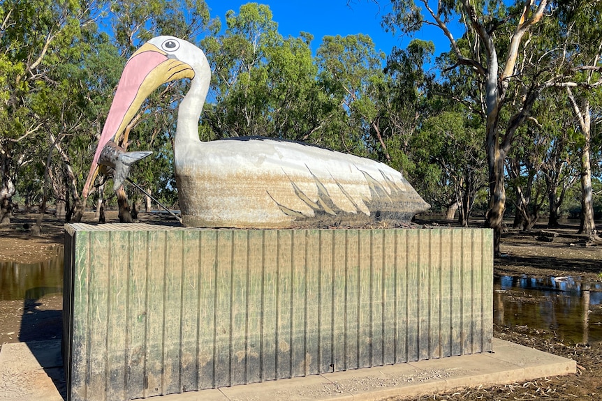 A giant pelican statue on a water damaged slab, blue skies, trees and low-lying water surrounding it. 
