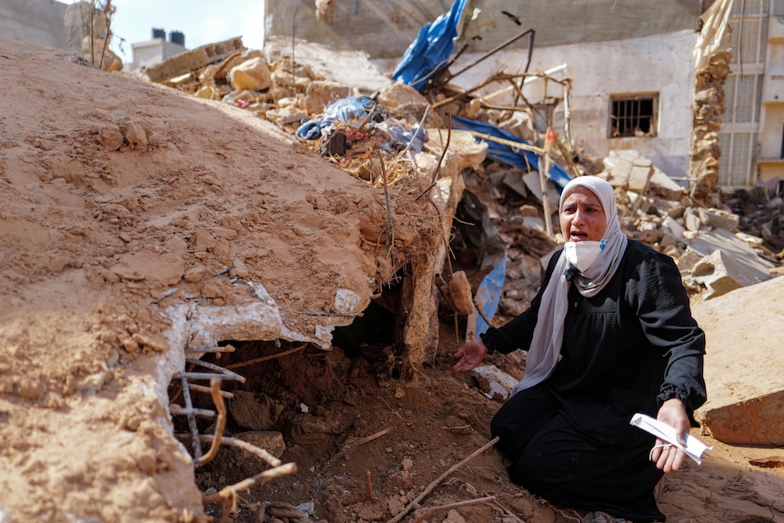 A woman kneels down in front of rubble and spreads her arms, her mouth open with a sad expression