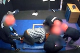 Several adults surround and hold down a boy in a padded room. The adults are wearing helmets.