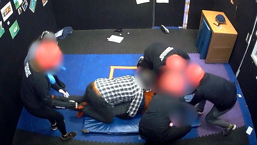 Several adults surround and hold down a boy in a padded room. The adults are wearing helmets.
