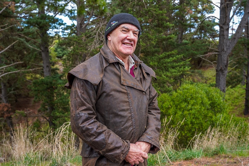 A man wearing a beanie and outdoor brown raincoat stands in a forested area smiling.