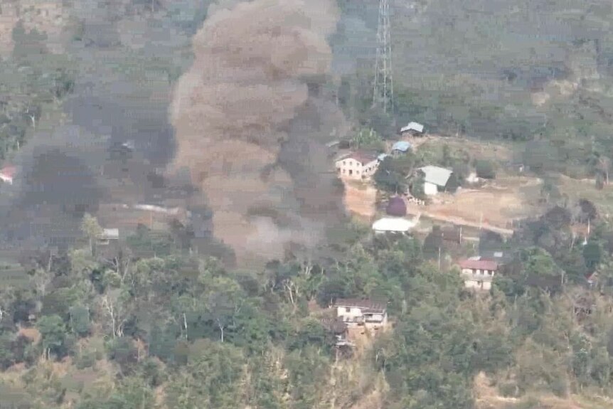 An aerial photo shows a plume of grey smoke rising from a small settlement surrounded by vegetation.