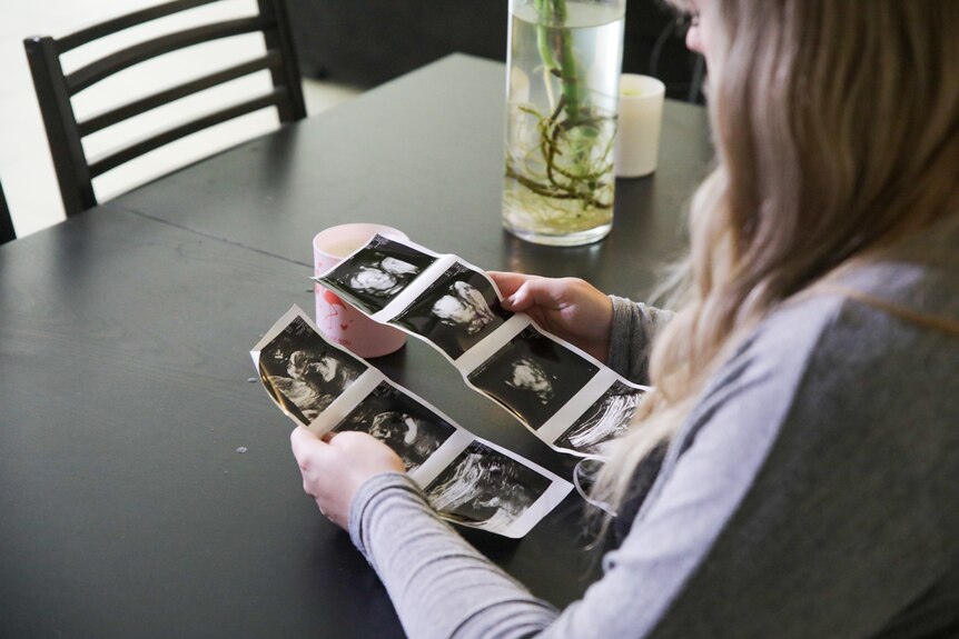 A close-up shot from over the shoulder of a young woman with blonde hair sitting at a table looking at baby ultrasound pictures.