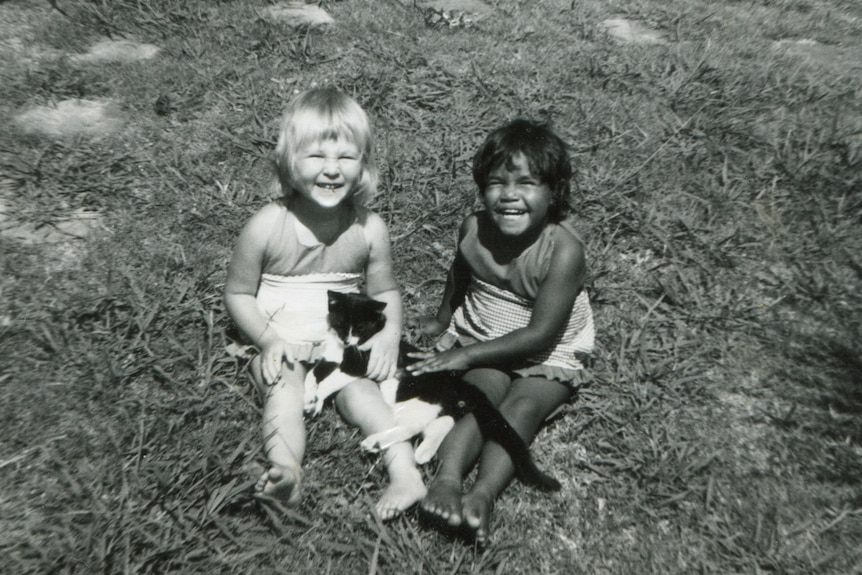 Two young girls, one white and one black, sit on the grass together smiling at the camera