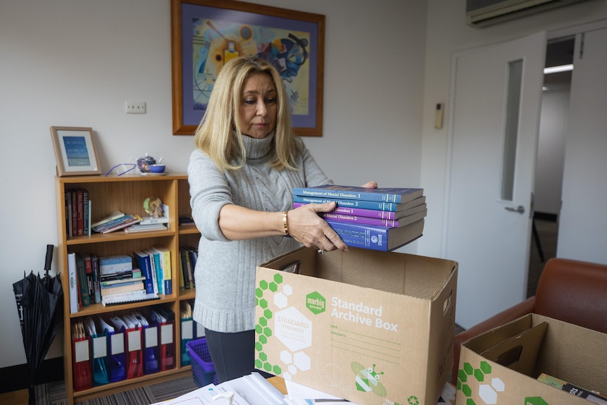 Woman in a grey polo neck puts books into a box as she packs up her office 