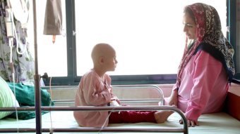 A child attached to a drip sits on a hospital bed with her mother.