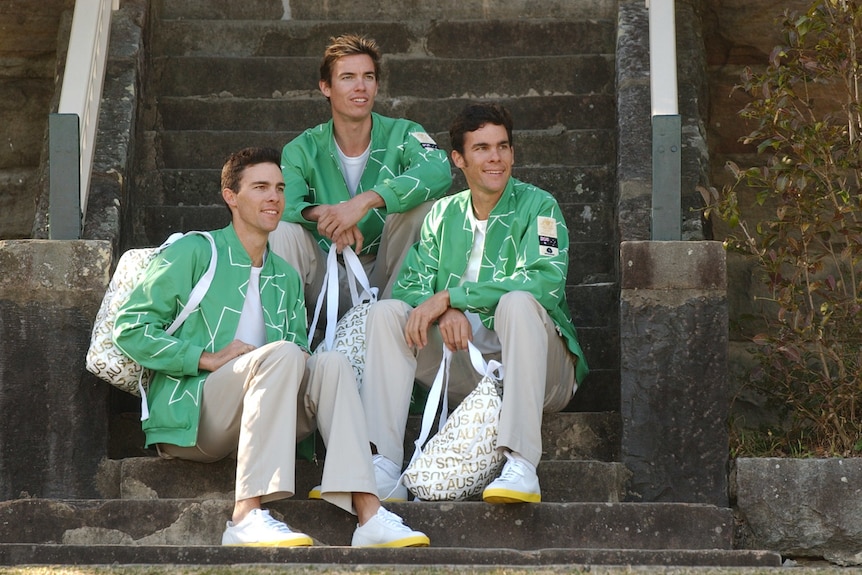 Three male athletes sit on steps wearing grey trousers and green jackets with white outlines 