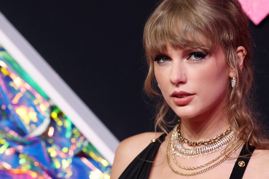 A close up of Taylor Swift at an event, where her wavy hair is partly pulled back and she wears several gold necklaces.