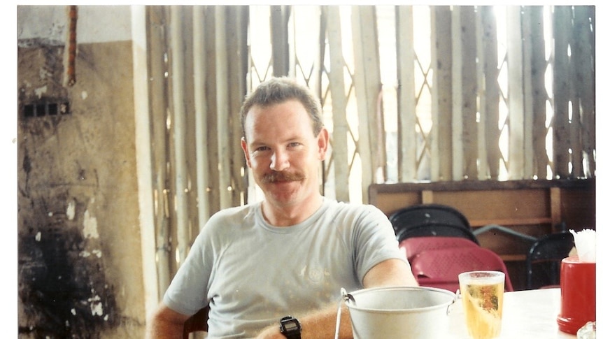 Mick Quinn in a sun-filled hut sits by a table with a cold drink on it. He smiles at the camera.