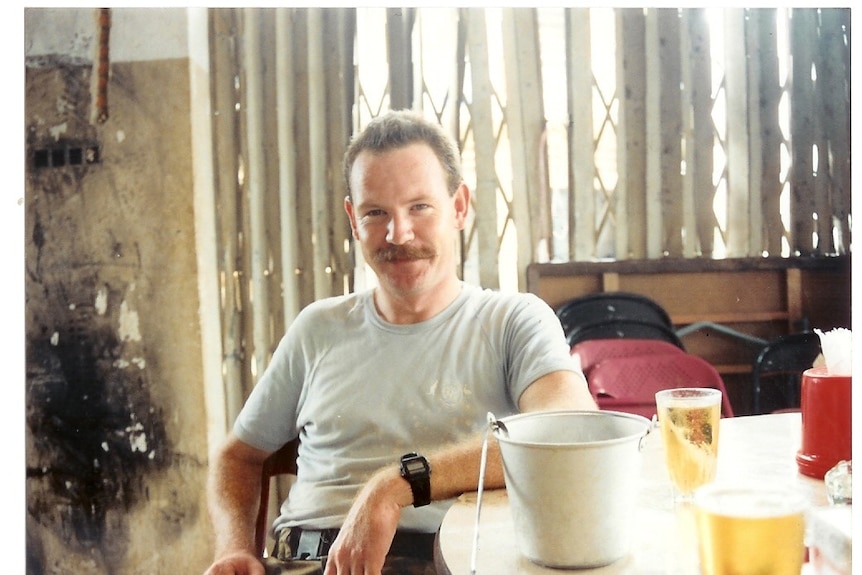 Mick Quinn in a sun-filled hut sits by a table with a cold drink on it. He smiles at the camera.