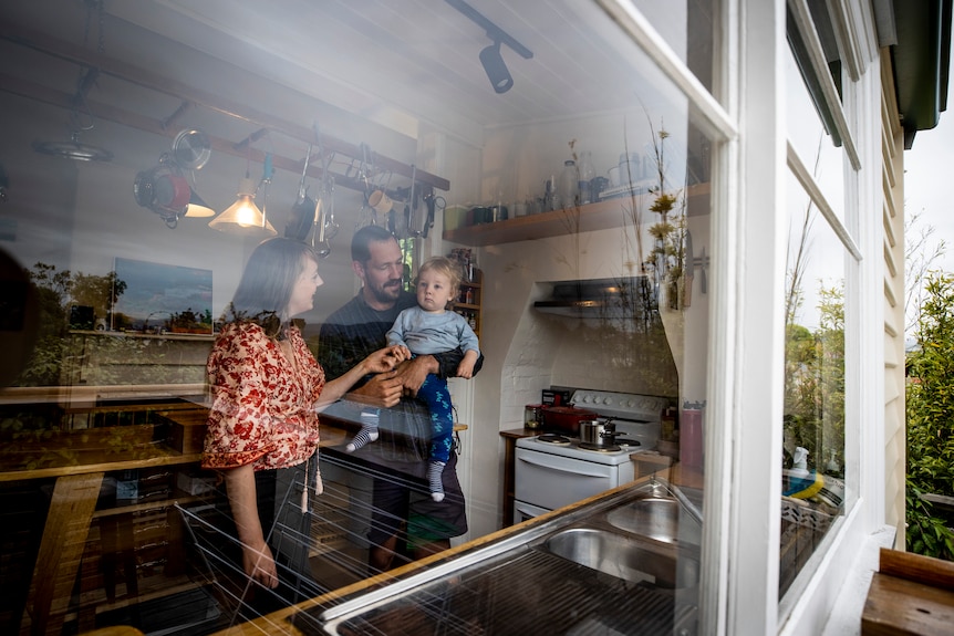 Una madre y un padre juegan con su bebé.  La foto fue tomada desde afuera a través de la ventana de su cocina.