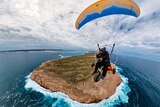 A man paramotoring high up in the air, blue sea is underneath him and a rugged island.