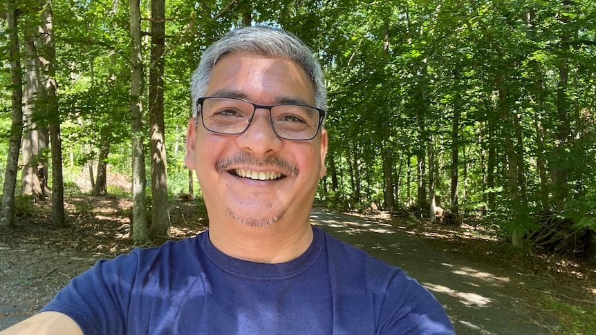 A man with grey hair and glasses smiling with lush green trees in the background. 