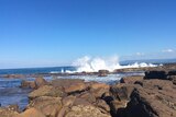A wave crashes against a rock platform on a sunny day.