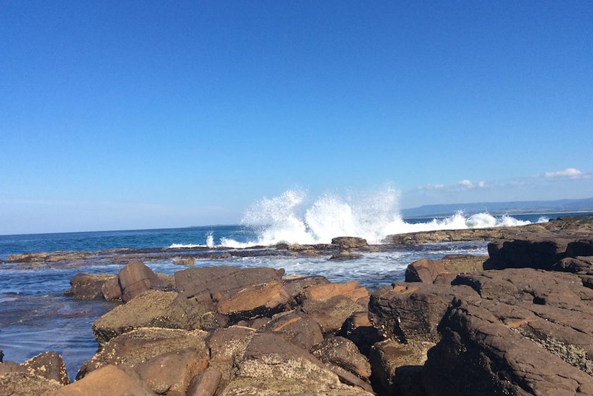 A wave crashes against a rock platform on a sunny day.