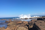 A wave crashes against a rock platform on a sunny day.