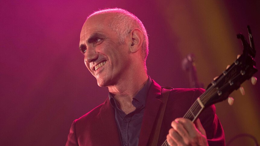 Paul Kelly is smiling and holding a guitar on stage in front of a pink and orange background
