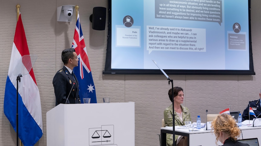 A man stands at a lectern gesturing to an image projected on the wall of a transcript of a phone conversation involving Putin