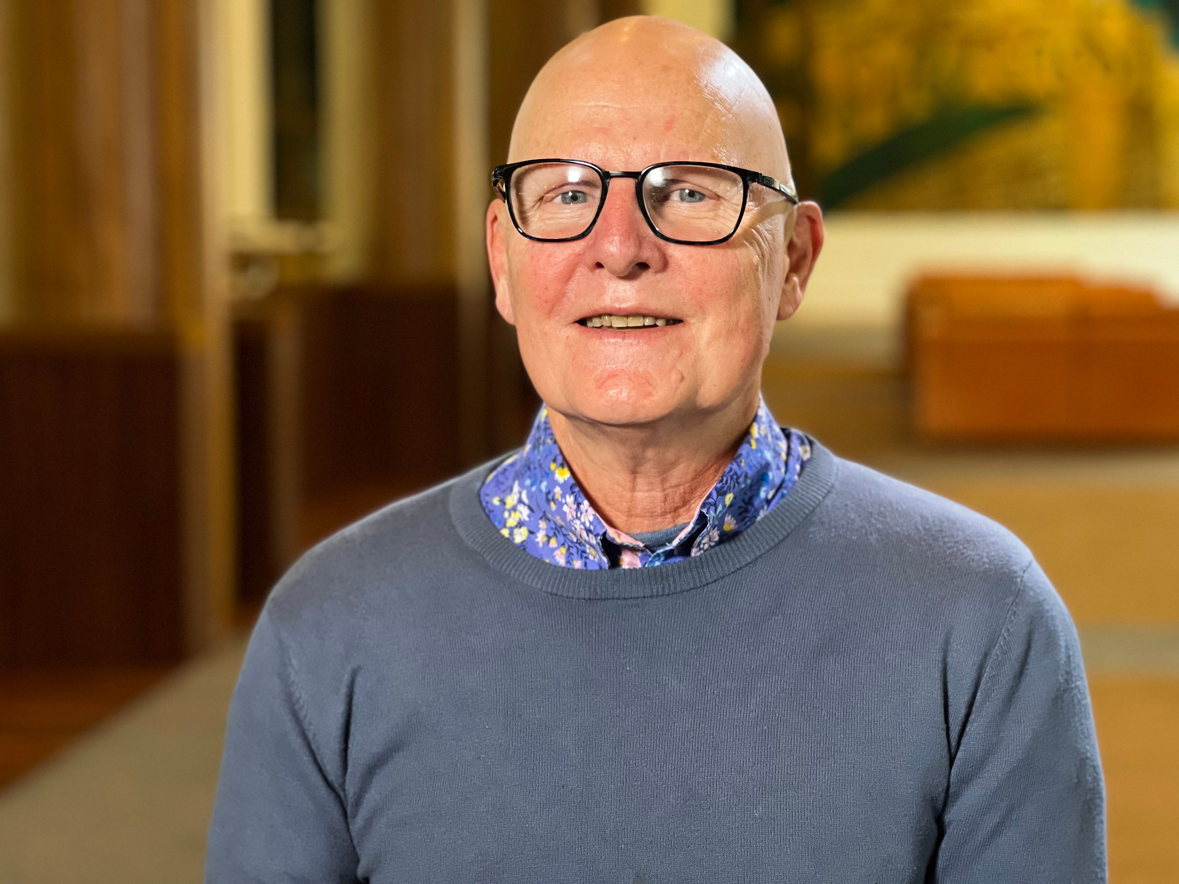 A man wearing a light grey jumper and glasses looks at the camera with a slight smile. 