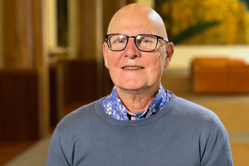 A man wearing a light grey jumper and glasses looks at the camera with a slight smile. 