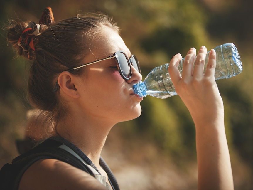 A young woman wearing sunglasses outside finishes the last sip of a water bottle.