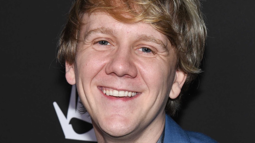 Australian comedian Josh Thomas stands in front of a media banner smiling wearing a blue jacket