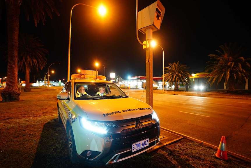 Two men sit in a car next to a red light camera in preparation to test it at an intersection