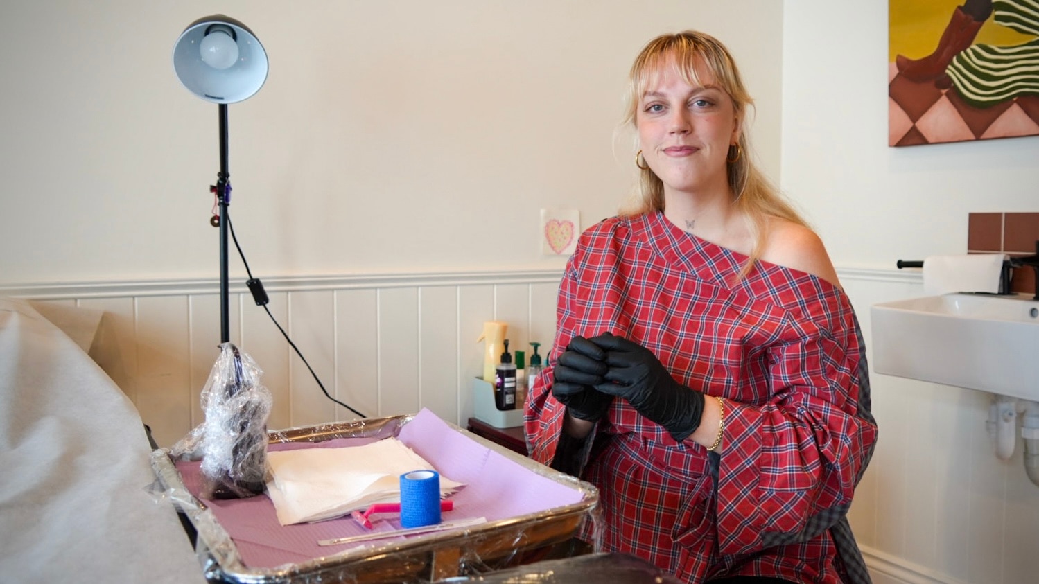 Blonde woman wearing a red checkered top with tattoo tray set up in front of her, putting on gloves and smiling at camera
