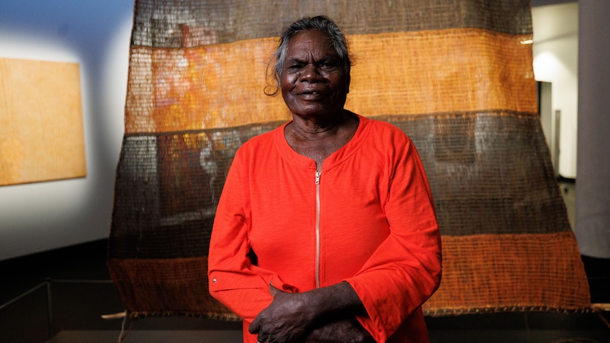 A smiling woman wearing a bright orange jacket standing in front of a large thatched artwork, inside an art gallery.