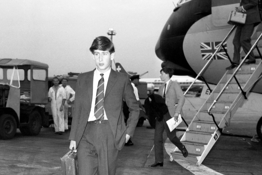 Prince Charles holding a suitcase and wearing a charcoal colored suit and school tie