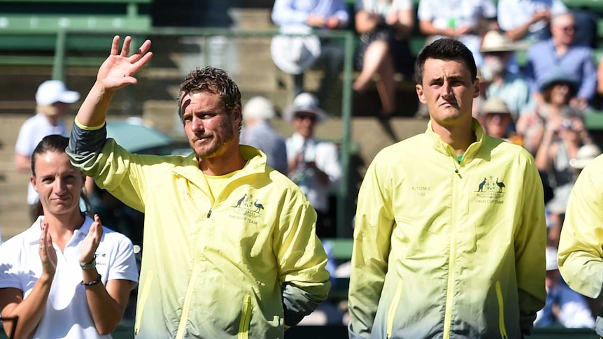 Lleyton Hewitt waves while standing next to Bernard Tomic. Both are looking into the sun, wearing Australian team colours.