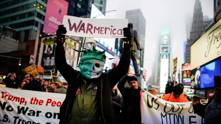 A man wearing a skull mask and a sign that says 'WARmerica' holds up a sign at a protest.