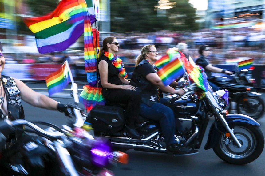 Two women ride a black motorbike along a parade route filled with crowds, there are lots of rainbow flags attached to the bike.