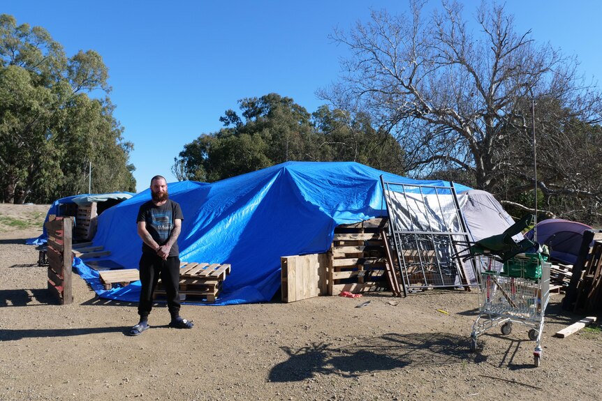 A man stands in front of a large tarp and a few tents propped up by pallets.