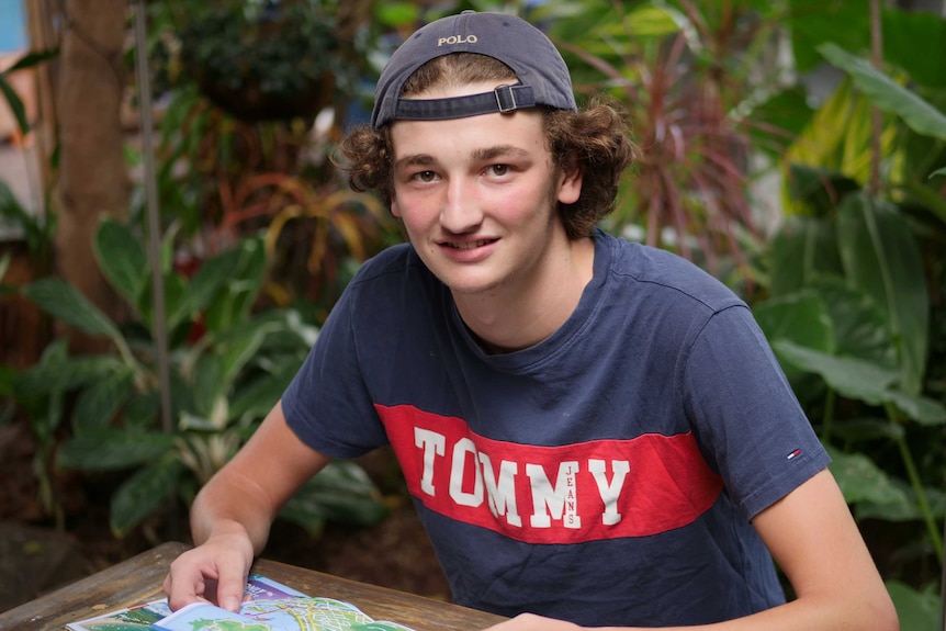 A young fellow with curly hair, wearing his cap backwards, sits at an outdoor table holding a map.