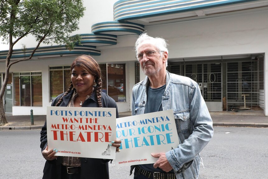 Marcia Hines and John Waters stand in front of a building with posters saying it should be a theatre