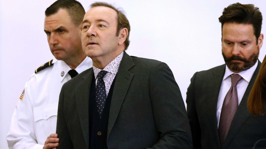 Actor Kevin Spacey enters the courtroom accompanied by a uniformed guard.