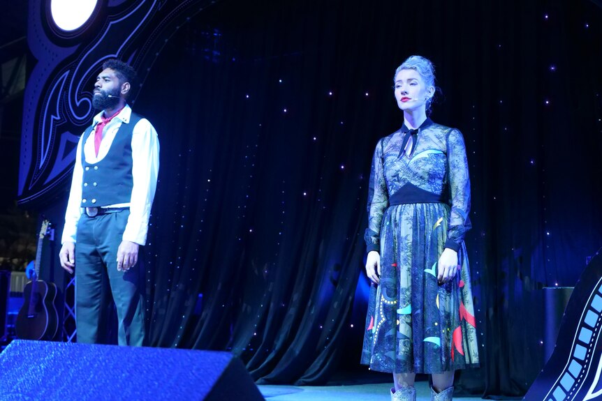 A man and woman stand on stage solemnly. There is a blue light. He is wearing a tuxedo top and jeans, she is wearing a dress.
