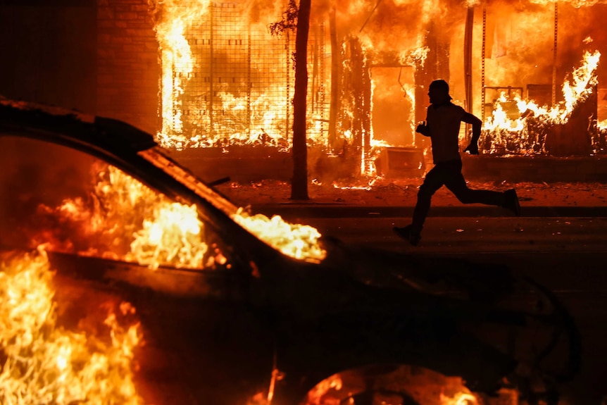 The outline of a man is seen as he runs between a burning building and a car set ablaze.