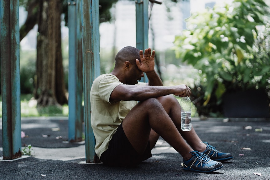 A man in a t-shirt sits on the ground with a bottle of water, wiping his sweaty brow after exercising