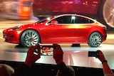 A red car on a stage in front of a crowd taking photos on their smart phones.