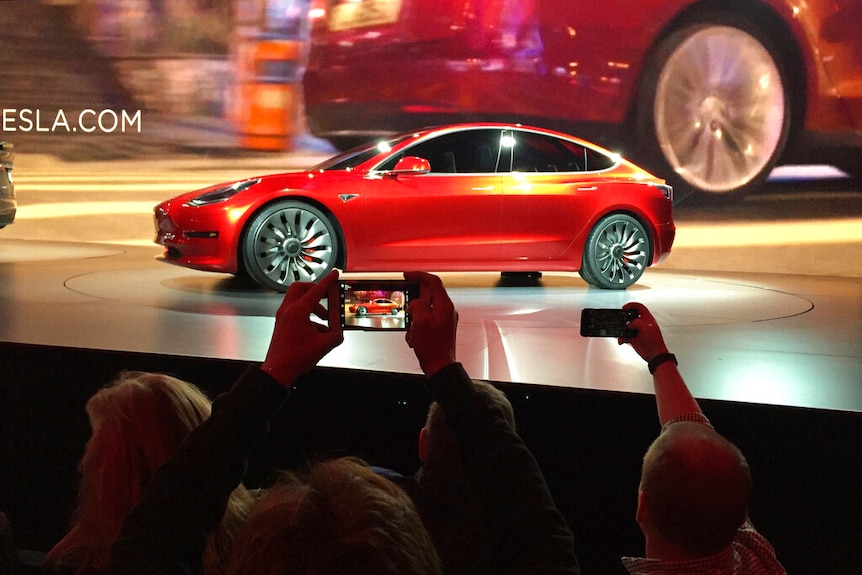 A red car on a stage in front of a crowd taking photos on their smart phones.