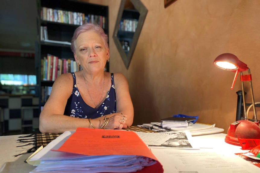 A woman with white hair, in blue top sits at desk, surrounded by files. She looks sad and stares off camera.