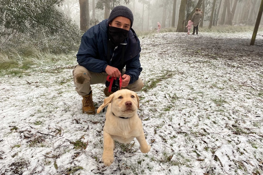 A man dressed in snow gear and his puppy crouch down in the snow.