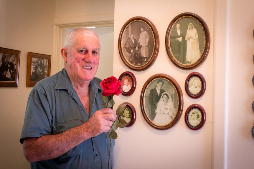 Monash engineer Neil Webber stands next to photographs of his parents and family while holding a single red rose.