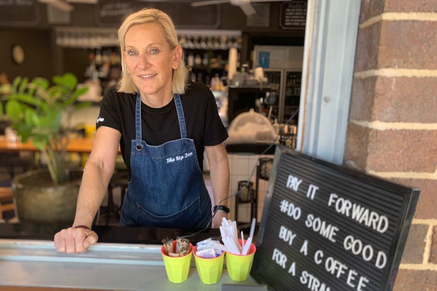 A woman in a black tshirt and denim apron stands behind a cafe counter.